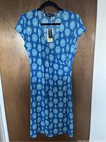 Front view of a blue women's dress with a white and light blue floral pattern, short sleeves, and V-neck, hanging on black hanger against wood background. Tag attached.