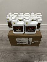 Full set of 12 jugs on top of the shipping box and 8 more jugs behind them, total of 20 jugs shown, all white plastic containers with the same label