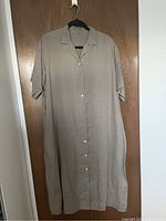 Front view of new Muji women's linen dress, beige color, button-down, mid-length sleeves, hanging on black hanger against wooden door.