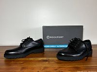 Pair of black men's leather shoes displayed in front of the Rockport shoe box, showing the shoes from the side and top angle.