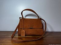 Front view of brown suede handbag with top handle and crossbody strap on wooden surface.