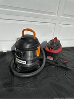 Photo showing two wet/dry vacuums, Shop-Vac with stainless steel tank and Craftsman smaller unit next to each other on black tarp