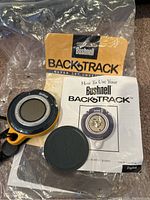 Bushnell BackTrack GPS device with black lanyard, protective cover, instruction manual, and original packaging on carpeted surface.