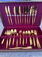Top view of the gold flatware neatly arranged inside the burgundy lined wooden case, showing knives, forks, spoons, and serving utensils.