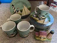 Set of plates and mugs stacked showing lighthouse and sailboat coastal design and two small lighthouse-themed collectibles