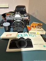 Front view of Minolta XG-1 with lens, leather case, film canister, manual, and filters on table