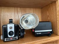 Shelf display showing Kodak Brownie Hawkeye Flash camera with clear flash attachment and Polaroid Sun 600 LMS instant camera