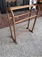 Wooden quilt rack standing on carpet in front of brick fireplace, showing side angle with three horizontal bars and rounded top sides.