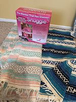 New pink Snuggie blanket in original box displayed with two colorful woven blankets from Mexico on the floor.