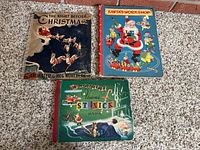 Three vintage Christmas pop up books laid on carpet, titles visible: 'The Night Before Christmas,' 'Santa's Workshop,' and 'St. Nick Magic.'