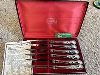 Full view of the 6 stainless steel cutters with ornate handles inside original red box with satin lining, including Carvel Hall guarantee certificate and Christmas card.