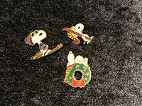 Top view of three Snoopy enamel pins on black fabric, showing colorful designs
