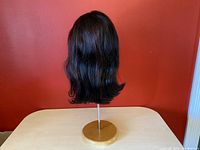 Photo showing back view of the dark brunette bob wig with slight waves at the ends on a wooden stand against an orange background.
