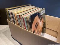 Box of 50 vinyl record albums showing spines and album covers stacked upright inside a cardboard box.