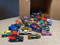 Overview photo of about 40-50 colorful die cast cars including sports cars, vintage cars, and fantasy car models all spilled in front of a box showing large lot