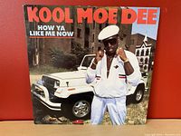 Front cover of Kool Moe Dee's How Ya Like Me Now album showing artist in white suit and cap with white jeep and brick buildings behind.