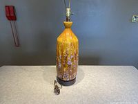 Front view of a tall ceramic lamp with fat lava glaze in yellow, brown and white dripping pattern, placed on a countertop against a gray background.