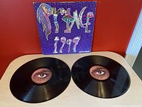 Two black vinyl records laid flat showing their labeled centers with eye graphics and text, with the purple album cover behind them showing 'Prince 1999' text.
