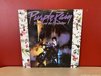 Front cover of the Purple Rain vinyl album featuring Prince on a motorcycle with smoky street background and floral border edges.