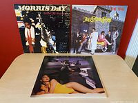 Three vintage vinyl record albums by Morris Day and The Time displayed upright and flat on a light-colored wooden table against a red background.