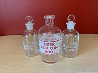 Photo of three vintage glass chemistry bottles: central larger Pyrex bottle labeled Nitric Acid Con. with two smaller Wheaton ammonium hydroxide bottles with round glass stoppers on either side, shown against a red background on a wooden surface.