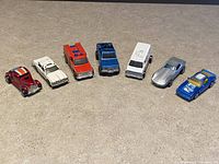 Wide shot showing all 7 vintage Hot Wheels cars in a row on a beige carpet surface.