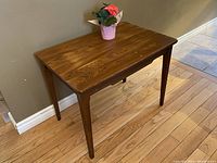 Full view of the teak wood vintage side table with tapered legs, placed against a wall and hardwood floor.