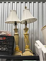 Two gold-toned table lamps with cream fabric shades sitting side by side, cords wrapped around bases, photographed against a corrugated metal background.