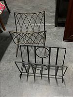 Two metal magazine racks on concrete floor, one with gold wire crisscross pattern and one black with vertical bars and circles.