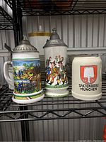 3 beer steins on a black metal rack; two with pewter lids showing German scenes, third is ceramic mug with Spatenbrau Munchen logo