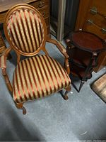 Front and side view of wooden accent chair with oval back and striped red and gold upholstery next to wooden side table with scalloped carrier-style top.