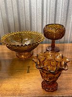 Three amber glass decorative pieces on wooden surface against corrugated metal background: a large hobnail footed bowl with ruffled edge, a pedestal compote, and a smaller hobnail ruffled vase.