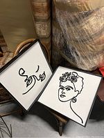 Two framed square modern line art pieces set leaned against a chair and stacked books.