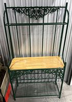 Front view of green metal baker's rack with wooden middle shelf and decorative scrollwork, against a corrugated metal background.