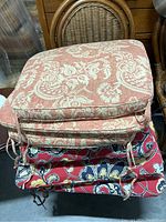 Stack of six chair cushions showing red with white floral paisley patterns and red background floral cushions.