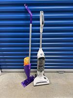 Shark vacuum and Swiffer Wet Jet mop shown side by side on concrete floor against blue door.
