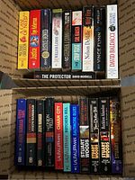 Photo shows two cardboard boxes filled with crime and suspense paperback books arranged vertically and stacked, revealing various titles and authors.