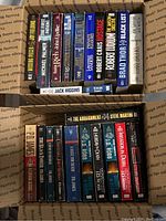 Top-down view of 11 crime and suspense paperback books arranged side-by-side in a box, showing titles and author names such as Robert Crais, Brad Thor, Michael Palmer, Jack Higgins.
