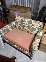 Loveseat with floral upholstery and pink seat cushion showing wear, stains, and storage surroundings.