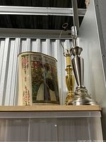 Photo showing two metal lamp bases (one silver, one gold-tone) next to a decorative cylindrical metal trash can with vintage printed design.