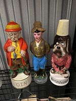 Three ceramic liquor decanters in character shapes: elderly man with pipe 'GIN', woman in traditional clothing, dog with lampshade hat 'Brights of Dog's Life'