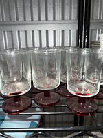 Front angle view of eight clear glass liquor drinking glasses with red stems lined up on a shelf
