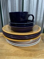 Stack of mixed dinnerware including plates in mustard yellow, deep purple, white, and a dark blue cup and saucer on top, showing the range of items included.