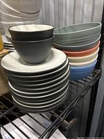Stack of white Noritake plates with two black bowls on top, placed on a metal rack with plastic bowls stacked nearby.