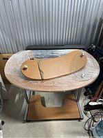 Desk with visible oval wooden top, metal frame, lower wooden shelf, curved glass shelf with metal standoffs, and broken wooden glass support piece.