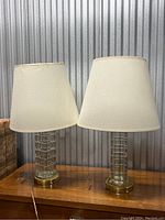 Full view of the pair of mid-century modern style crystal lamps with cream fabric shades placed on a wooden surface against a corrugated metal background.