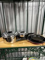 Set of kitchen pots and pans on a wooden shelf against a metallic background. Includes one large stainless steel pot with lid, a smaller pot with lid and black handle, and two black frying pans.