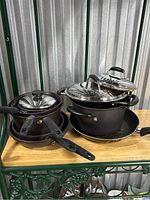 Full view of six black Silverstone pots and pans on wooden shelf, some with glass lids on top.