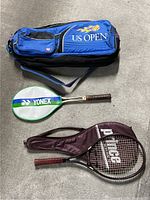 Photo of two tennis rackets with their protective covers and a US Open blue racket bag on a concrete floor.