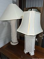 Two white lamps on a wooden surface next to a mirror. One cylindrical with leaf designs, the other rectangular with textured pattern, both with white fabric lampshades.
